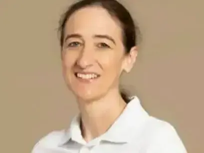 Eine lächelnde Frau mit kurzen Haaren, die ein weißes Polohemd trägt, steht vor einem beigen Hintergrund.