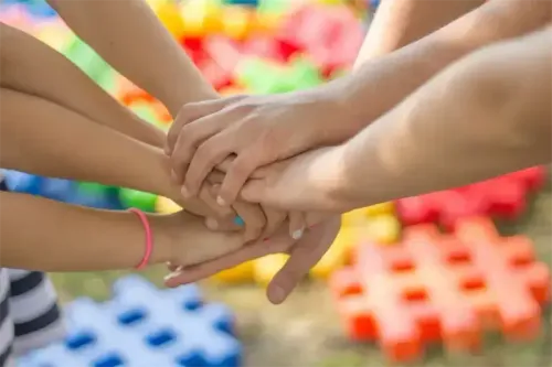 Mehrere Kinder halten sich in einer kreisförmigen Formation an den Händen. Sie befinden sich auf einem Spielplatz mit bunten Spielzeugen im Hintergrund.