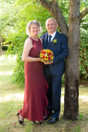 Ein Mann und eine Frau stehen neben einem Baum und lächeln für ein Foto. Der Mann trägt einen Anzug und eine Krawatte, und die Frau trägt ein Weinrotkleid. Der Mann hält einen Blumenstrauß.