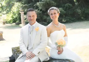 Ein Mann und eine Frau in Hochzeitskleidung sitzen auf einer Steinbank in einem Garten. Der Mann trägt einen weißen Anzug mit einer gelben Blume an der Revers, während die Frau ein weißes Hochzeitskleid trägt und einen Blumenstrauß hält.