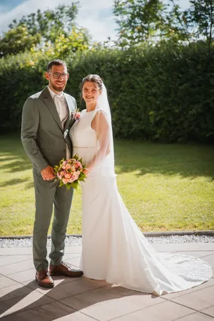 Ein Mann und eine Frau stehen zusammen auf einer Terrasse und lächeln für ein Foto. Die Frau trägt ein weißes Hochzeitskleid und einen Schleier, und der Mann trägt einen grauen Anzug und eine Brille. Sie sind von grünem Gras und Büschen umgeben.