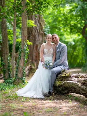 Ein Brautpaar sitzt auf einem Baumstamm im Wald und lächelt in die Kamera. Die Braut trägt ein weißes Hochzeitskleid und hält einen Blumenstrauß.