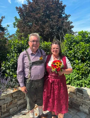 Ein älterer Mann und eine Frau stehen lächelnd vor einer Steinmauer. Der Mann trägt ein kariertes Hemd und Hosenträger, und die Frau trägt ein rotes Kleid und hält einen Blumenstrauß.