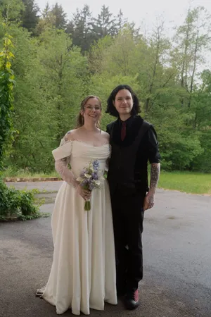 Eine Frau in einem Hochzeitskleid und ein Mann in einem schwarzen Anzug stehen auf einem Parkplatz mit einem Blumenstrauß.