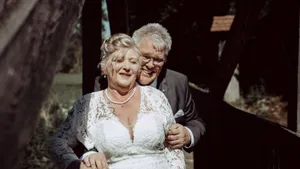 Ein älteres Paar lächelt und posiert für ein Foto vor einer Holzbrücke. Die Frau trägt ein weißes Hochzeitskleid und eine Halskette, während der Mann einen Anzug und eine Brille trägt.