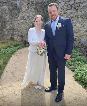 Ein Mann und eine Frau stehen lächelnd auf einem Pfad. Die Frau hält einen Blumenstrauß und trägt ein weißes Kleid. Der Mann trägt einen Anzug und eine Krawatte.