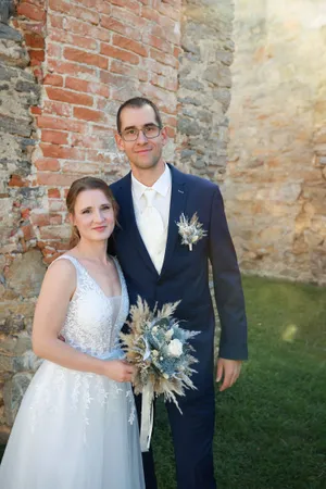 Ein frisch verheiratetes Paar steht eng beieinander, die Braut in einem weißen Kleid und der Bräutigam in einem blauen Anzug, vor einer Backsteinmauer.