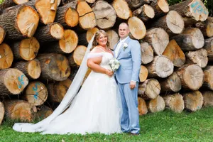 Ein frisch verheiratetes Paar steht vor einem großen Haufen Holzscheite und lächelt für ein Foto.