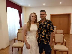 Ein Mann und eine Frau stehen zusammen in einem Raum mit Holzböden und weißen Wänden. Die Frau trägt ein weißes Kleid und der Mann trägt ein schwarzes Hemd mit orangefarbenen Drucken.