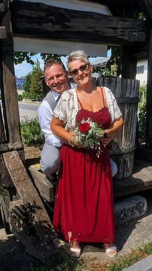 Ein Mann und eine Frau lächeln und posieren für ein Foto vor einer rustikalen Holzstruktur. Die Frau hält einen Blumenstrauß und trägt ein rotes Kleid.
