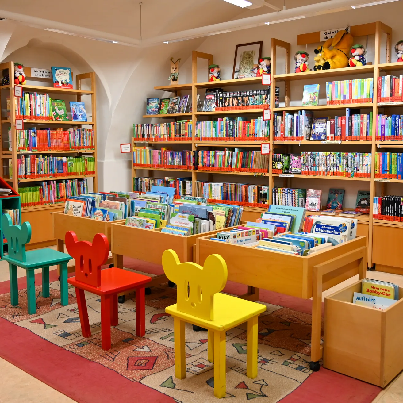 Eine Kinderbibliothek mit bunten Regalen und Büchern. Es gibt Micky-Maus-Stühle und einen Teppich auf dem Boden.