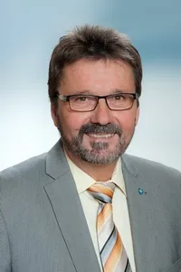 Ein Mann mit Brille und Bart, der einen grauen Anzug und einen gestreiften Krawatten trägt, lächelt für ein Foto.