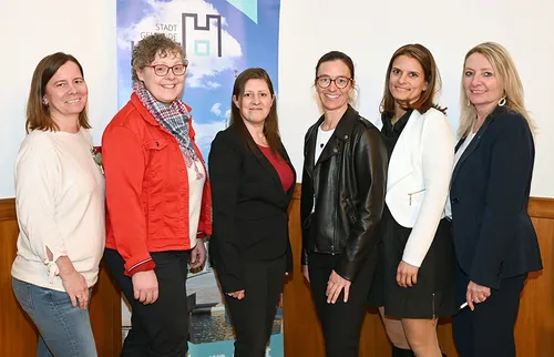Sechs Frauen in Berufskleidung stehen gemeinsam vor einem Hintergrund mit blauem Himmel und Wolken. Sie lächeln und posieren für ein Foto.