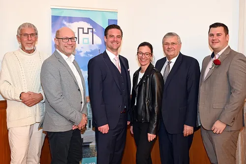 Fünf Erwachsene in formeller Kleidung stehen dicht beieinander vor einem Banner. Sie lächeln für die Kamera.