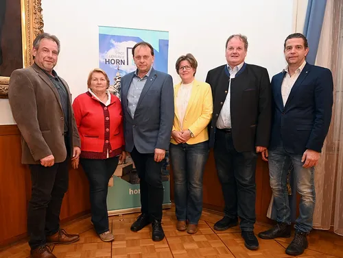 Eine Gruppe von sechs Erwachsenen steht vor einem Banner, das für die Stadt Horn wirbt. Sie alle lächeln und scheinen für ein Foto zu posieren.