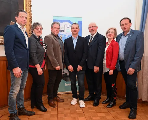 Sieben Menschen posieren für ein Foto drinnen, formell gekleidet, mit einem Banner hinter ihnen, das 'Stadt Endende' steht.
