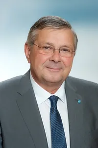 Ein lächelnder Mann in einem grauen Anzug und blauer Krawatte mit Brille. Er scheint sich in einer professionellen Umgebung zu befinden.