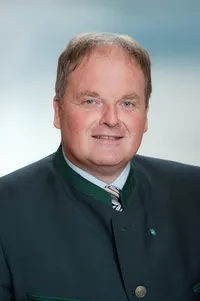 Ein Mann im Anzug mit einer grünen Weste lächelt für ein Porträt. Der Hintergrund ist hellblau.