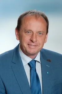 Ein Mann in einem blauen Anzug und einer Krawatte mit einer Anstecknadel, lächelt in die Kamera und steht vor einem blauen Himmel.
