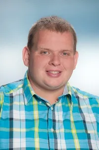 Ein Mann mit kurzen Haaren, der ein blau-weiß kariertes Polohemd trägt, lächelt in die Kamera. Er scheint sich in einer professionellen Umgebung zu befinden.