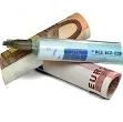 Ein Stapel gerollter Euro-Banknoten und eine Spritze auf weißem Hintergrund. Die Spritze ruht auf den Banknoten.