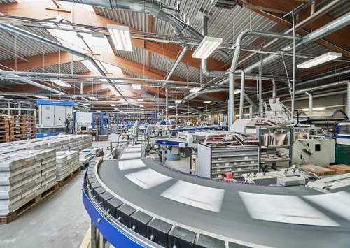 Eine große Fabrik mit einem gekrümmten Förderband, verschiedenen Maschinen und Lagerregalen. Die Decke ist aus Metall und es gibt Holzbalken.