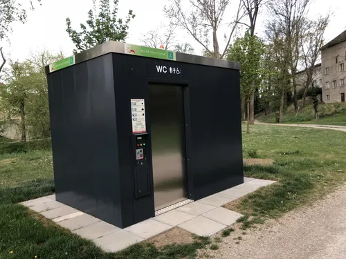 Eine schwarze WC-Toilettenkabine mit silberner Tür, Bedienfeld und offener Tür auf einer Betonplattform in einem Grasfeld.