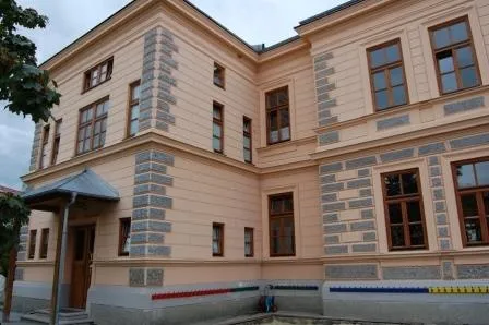 Ein großes beiges Gebäude mit mehreren Fenstern und einer grauen Steinfassade. Es verfügt über eine braune Eingangstür und einen grauen Vorbau. Das Gebäude hat ein modernes Design mit einer Mischung aus traditionellen und zeitgenössischen Elementen.