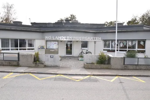 Ein weißes Gebäude mit einem Schild, das 'No Landeskindergarten' steht. Ein Fahrrad ist auf der Seite geparkt und davor stehen Topfpflanzen.