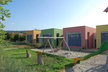 Ein Kinderspielplatz mit Schaukel, Rutsche und Bank, umgeben von bunten Häusern und Grün.