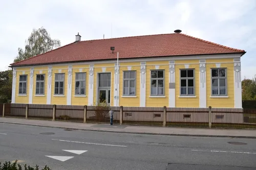 Ein gelbes Gebäude mit einem roten Dach und vielen Fenstern. Es hat einen Zaun vorn und einen Parkscheinautomat.