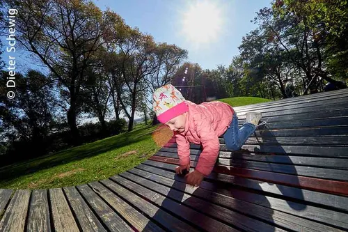 Ein Kind in einer rosa Jacke und Mütze klettert auf eine Holzbank im Park unter einem strahlenden Sonnenschein.