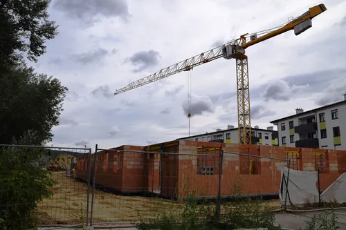 Eine Baustelle mit einem Kran und einem Gebäude im Bau. Das Gebäude ist aus Ziegeln und hat Fenster. Ein Zaun umgibt die Baustelle.