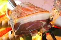 Ein Nahaufnahme eines Stücks gepökeltem Schinken, möglicherweise Prosciutto, auf einem Grill mit einem Feuer im Hintergrund. Der Schinken hat eine dünne Fettschicht und eine knusprige, goldbraune Kruste.