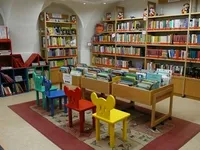 Ein Kinderbereich in einer Bibliothek mit bunten Stühlen und Regalen, die mit Büchern gefüllt sind. Die Stühle sind in einer Reihe angeordnet und die Regale ordentlich organisiert. Ein Teppich liegt auf dem Boden.
