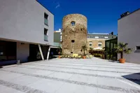 Ein leerer Innenhof mit einem großen Steinturm, modernen Gebäuden und mehreren Topfbäumen. Der Turm hat Fenster und eine Tür, und es gibt eine Terrasse mit Stühlen und Tischen.