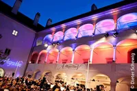 Ein Konzert unter Beleuchtung im historischen Innenhof. Das Publikum sitzt vor einem Orchester und wird von rosa und lila Lichtern beleuchtet.
