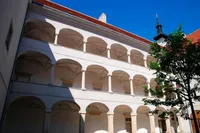 Ein klassischer europäischer Innenhof mit mehreren Bogenfenstern und einem roten Dach. Der Innenhof ist von hohen, weißen Säulen umgeben und mit einem Baum geschmückt.