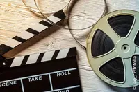 Ein Regisseursklatscher und eine Filmrolle liegen auf einer Holzoberfläche. Der Klatscher zeigt links 'Aufnahme' und rechts 'Roll', während die Filmrolle teilweise abgerollt ist.