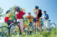 Eine Gruppe von Menschen steht mit ihren Fahrrädern auf einem Grasweg, einige tragen Helme und halten Wasserflaschen. Ein Individuum lächelt und spricht mit einem anderen.