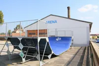 Eine große blaue Skateboard-Rampe steht vor einem weißen Gebäude mit der Aufschrift Arena. Die Rampe hat Graffiti und einen Metallzaun. Ein Schornstein befindet sich auf dem Dach des Gebäudes.