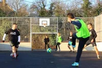 Eine Gruppe von Männern spielt draußen Fußball, einer kickt einen Ball in der Nähe eines Basketballfeldes. Sie tragen verschiedenfarbige Trikots und Sneakers.