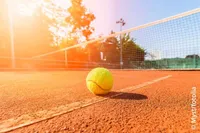 Ein Tennisball liegt auf einem Tennisplatz mit einem Netz im Hintergrund bei hellem Sonnenlicht.