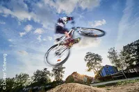 Eine Person fährt ein Fahrrad über einen Erdhügel, in der Luft, mit Häusern und Bäumen im Hintergrund unter einem blauen Himmel.