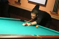 Ein Mann in Schwarz spielt Billard und zielt mit einem Queue. Der Billardtisch ist grün und zwei Bälle sind darauf, einer davon wird getroffen.