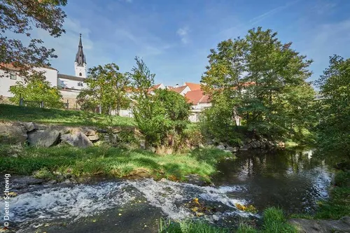 Eine ruhige Dorfszene mit einem Kirchturm, umgeben von Bäumen und einem fließenden Fluss unter einem blauen Himmel.