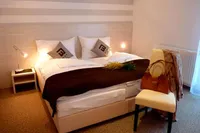 Ein Schlafzimmer mit einem Doppelbett, weißen Laken, brauner Decke und dekorativen Kissen. Eine Lampe auf einem Nachttisch und eine Handtasche auf einem Stuhl.
