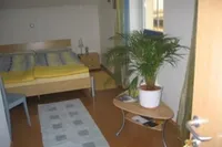 Ein Schlafzimmer mit einem Bett, einem Nachttisch und einer großen Topfpflanze. Ein Teppich liegt auf dem Boden in der Nähe eines Tisches. Der Raum hat weiße Wände und Holzböden.