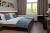Ein Schlafzimmer mit zwei Betten, jedes mit weißen Laken und blauen Decken, wird vom Sonnenlicht durch das Fenster beleuchtet. Die Wände sind weiß gestrichen und unter dem Fenster befindet sich ein Heizkörper.