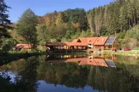 Eine rustikale Lodge mit rotem Dach und Solarpanels liegt am Rande eines ruhigen Sees, umgeben von üppigem Baumbestand und einem roten Auto.